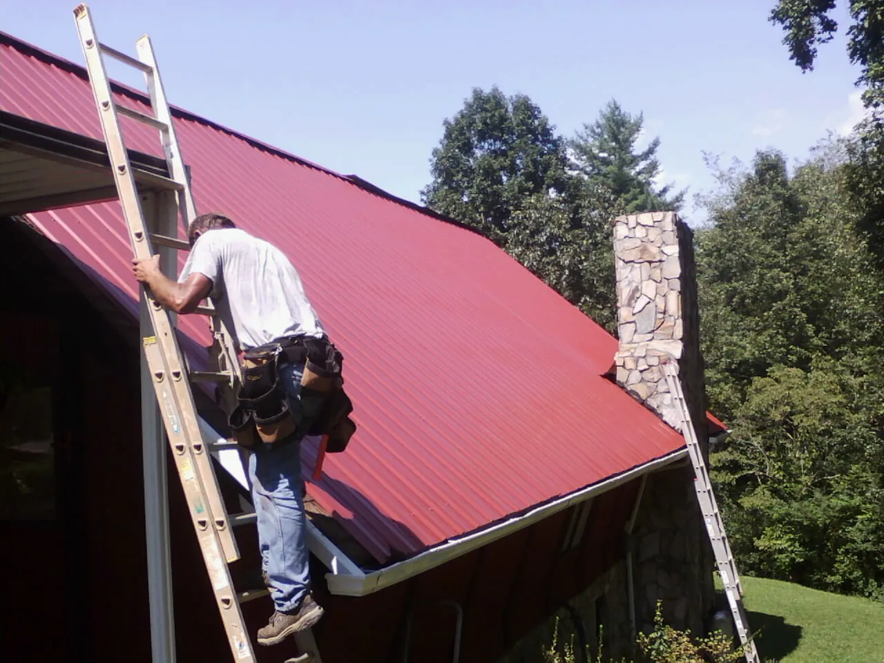 Roofer on a ladder beside a red metal roof and stone chimney — Soffit & Fascia Repair by Roofing Brothers in Clinton